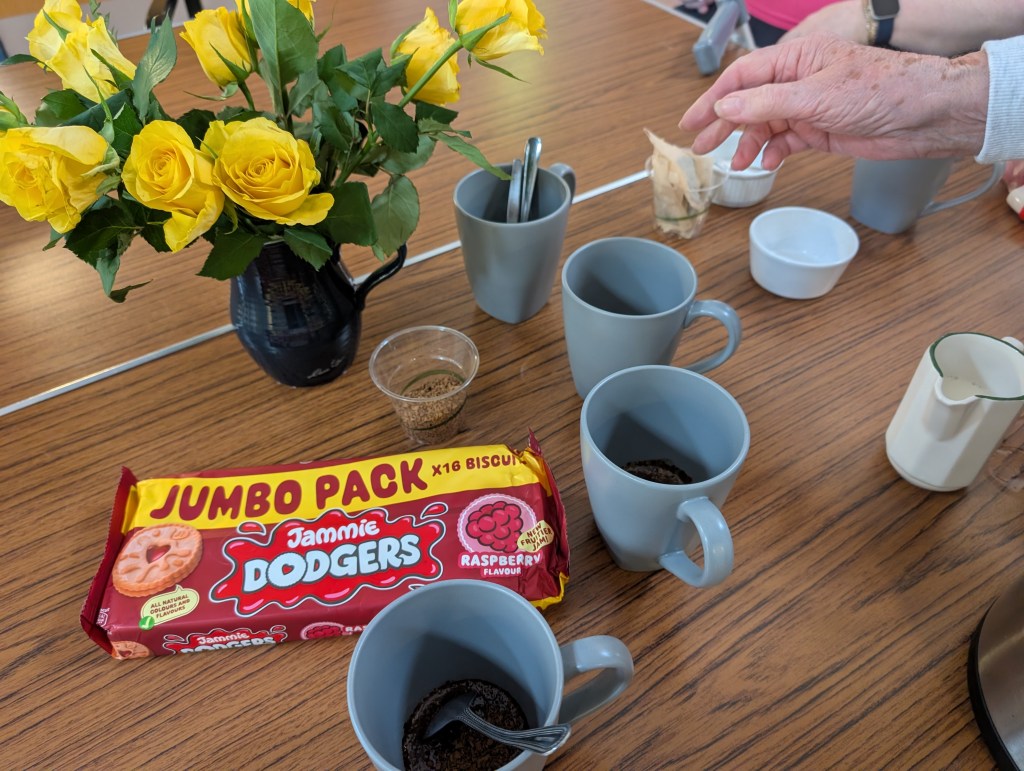 a table with some yellow roses in  vase, some empty mugs a jug of milk and bowl of sugar and a full packet of Jammy dodger biscuits. 